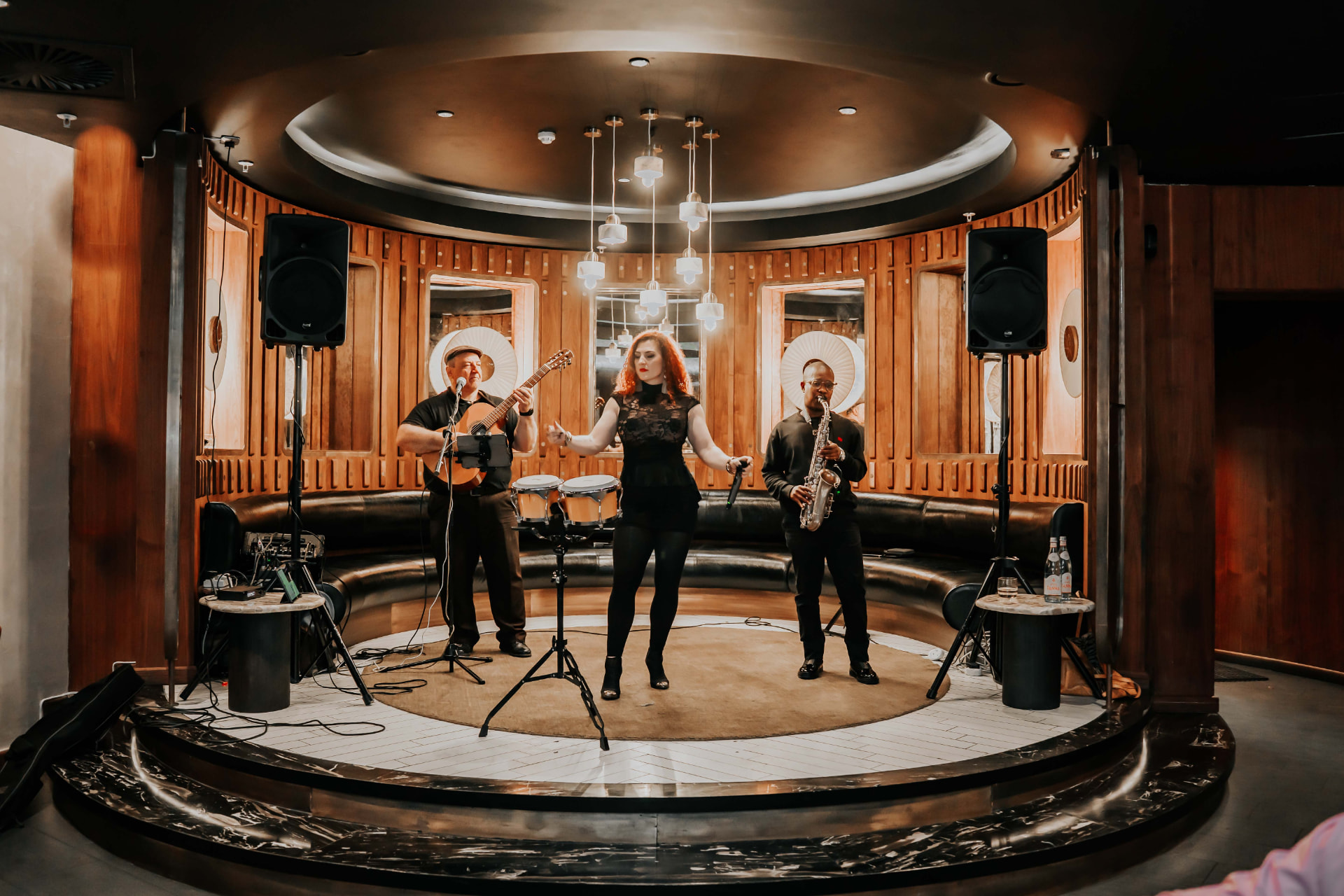 The full collective on a round stage in a timber-panelled room: guitarist, René centre with bongos, saxophonist.